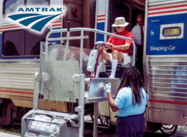 Logo: Amtrak. Staff members assist a person in a scooter as they exit the sleeping car of an Amtrak train onto a platform lift.
