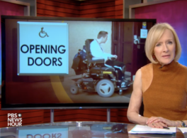 Screenshot from a PBS News Hour segment. The host speaks in front of a screen showing a person in a power wheelchair outside the door of a building with the caption "Opening Doors."